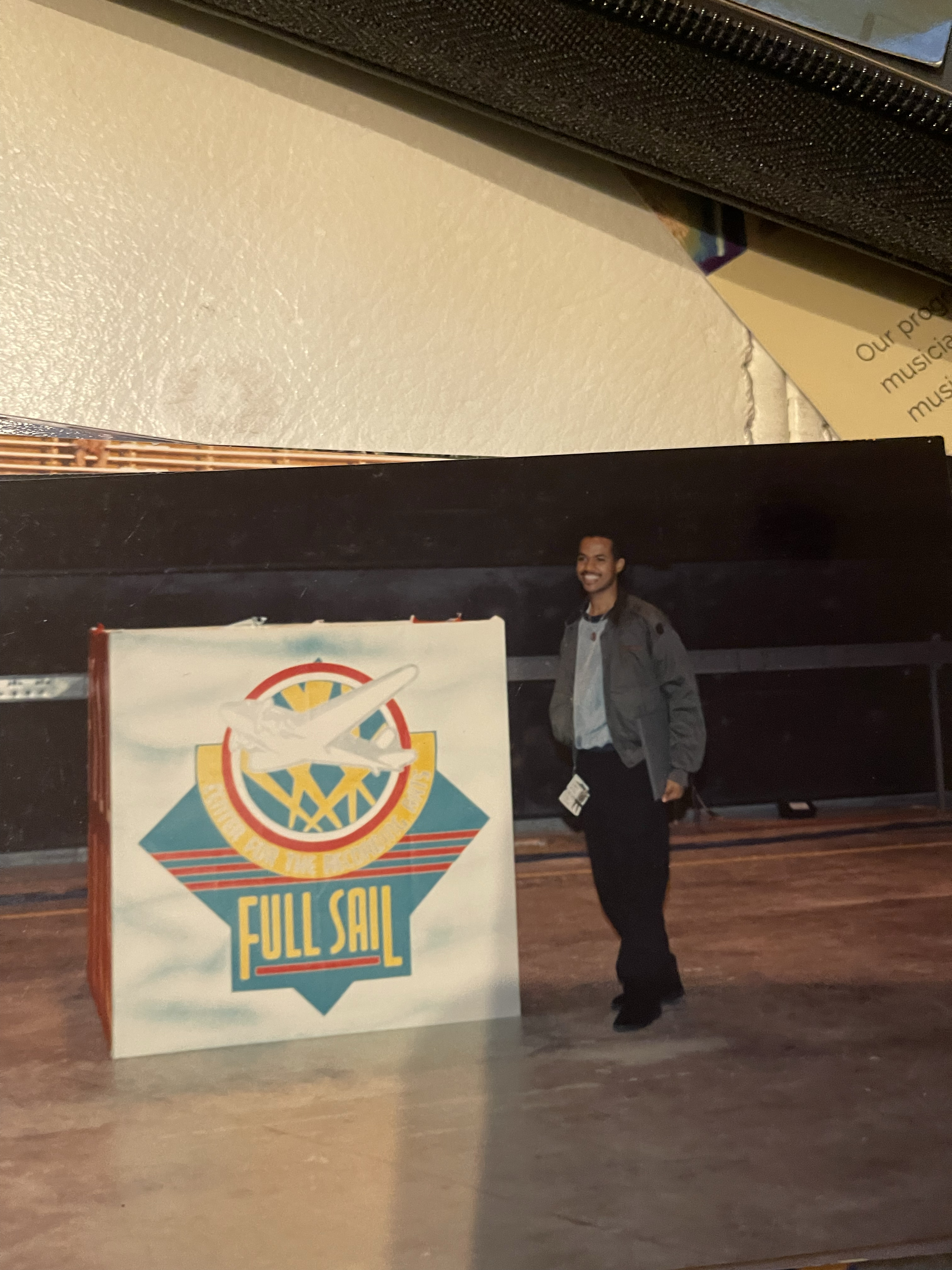 Byron Jupiter, Jr. smiling on a stage floor beside a Full Sail Center for the Recording Arts sign with the school's vintage airplane logo, Winter Park, Florida.