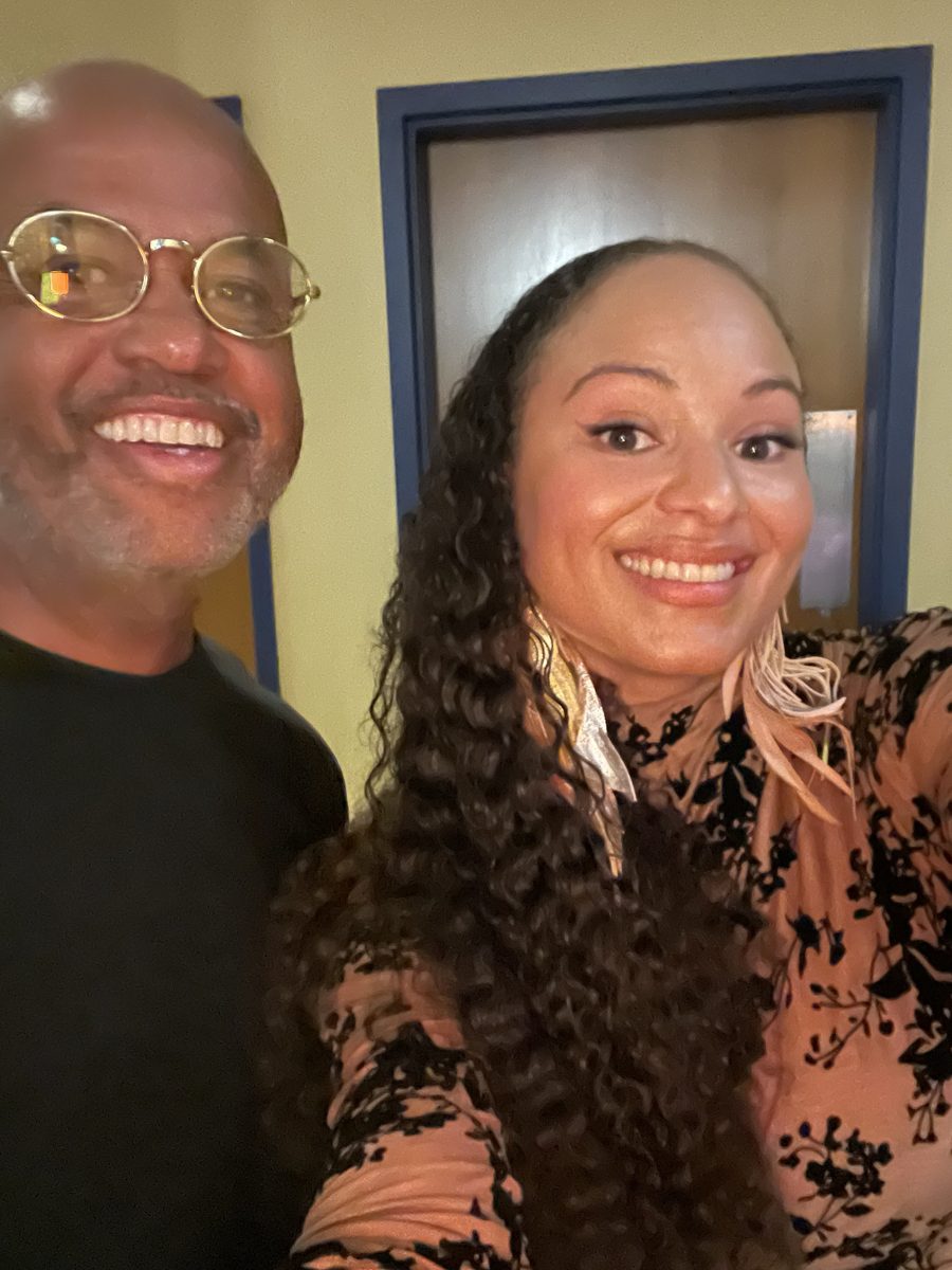 Selfie of two smiling New Orleanians: a man in gold-rimmed glasses and a black shirt beside a woman with long curly hair, feather earrings and a peach-and-black floral top.